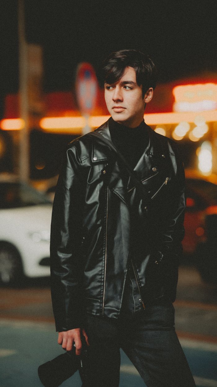 Fashionable man in a black leather jacket poses in an urban setting at night, exuding modern style.