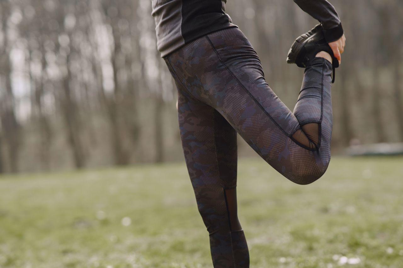 our-experience Side view anonymous crop fit female in dark leggings and black runners stretching legs in park before workout in daylight